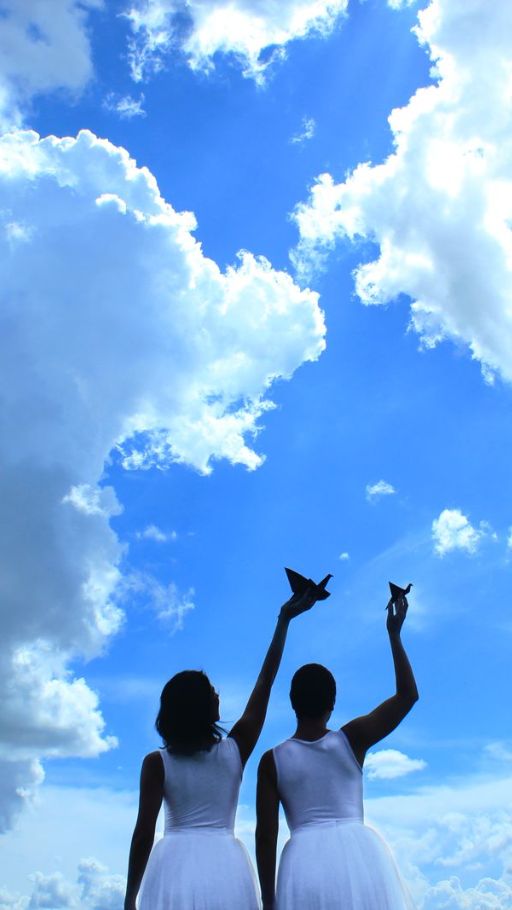 Imagem de divulgação do espetaculo Voa, com Cirila Targhetta e Tatiana Bittar. Um. ceu azul com nuvens compe o fundo.